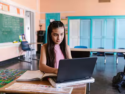Student using laptop and textbook at a desk in a classroom